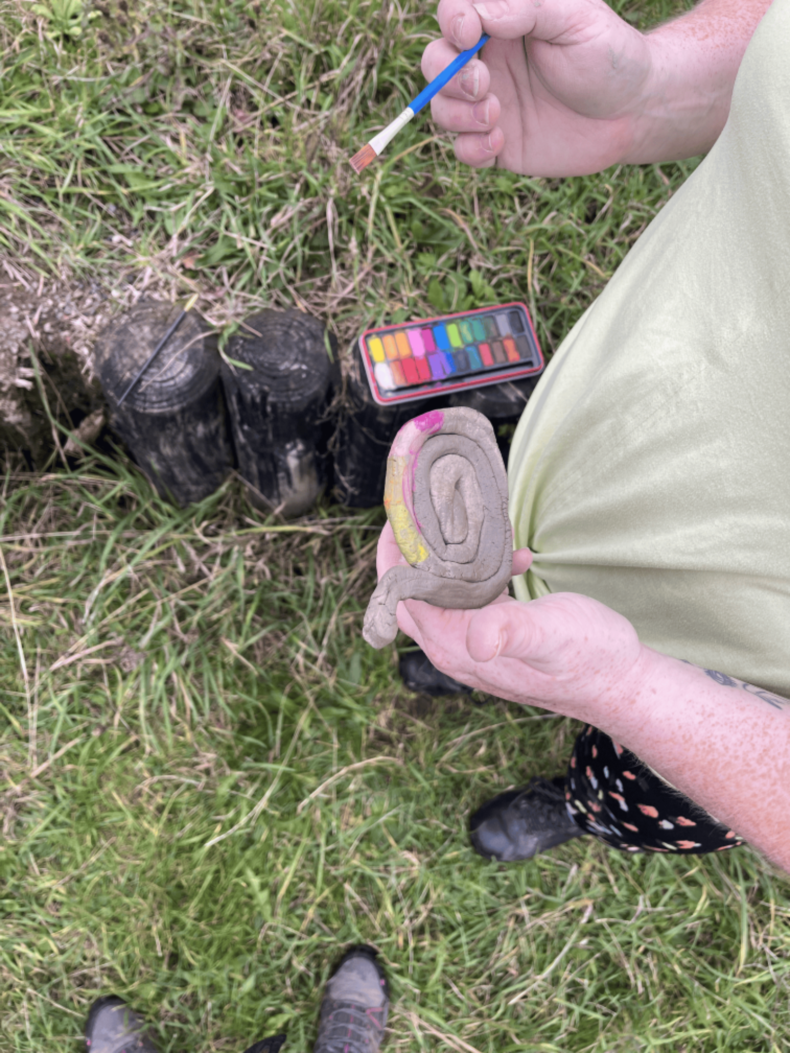 A person holds a spiral of grey clay in one hand and a pint brush in the other. They are stood in a grassy area and on a small fence sits a palette of colourful paints.