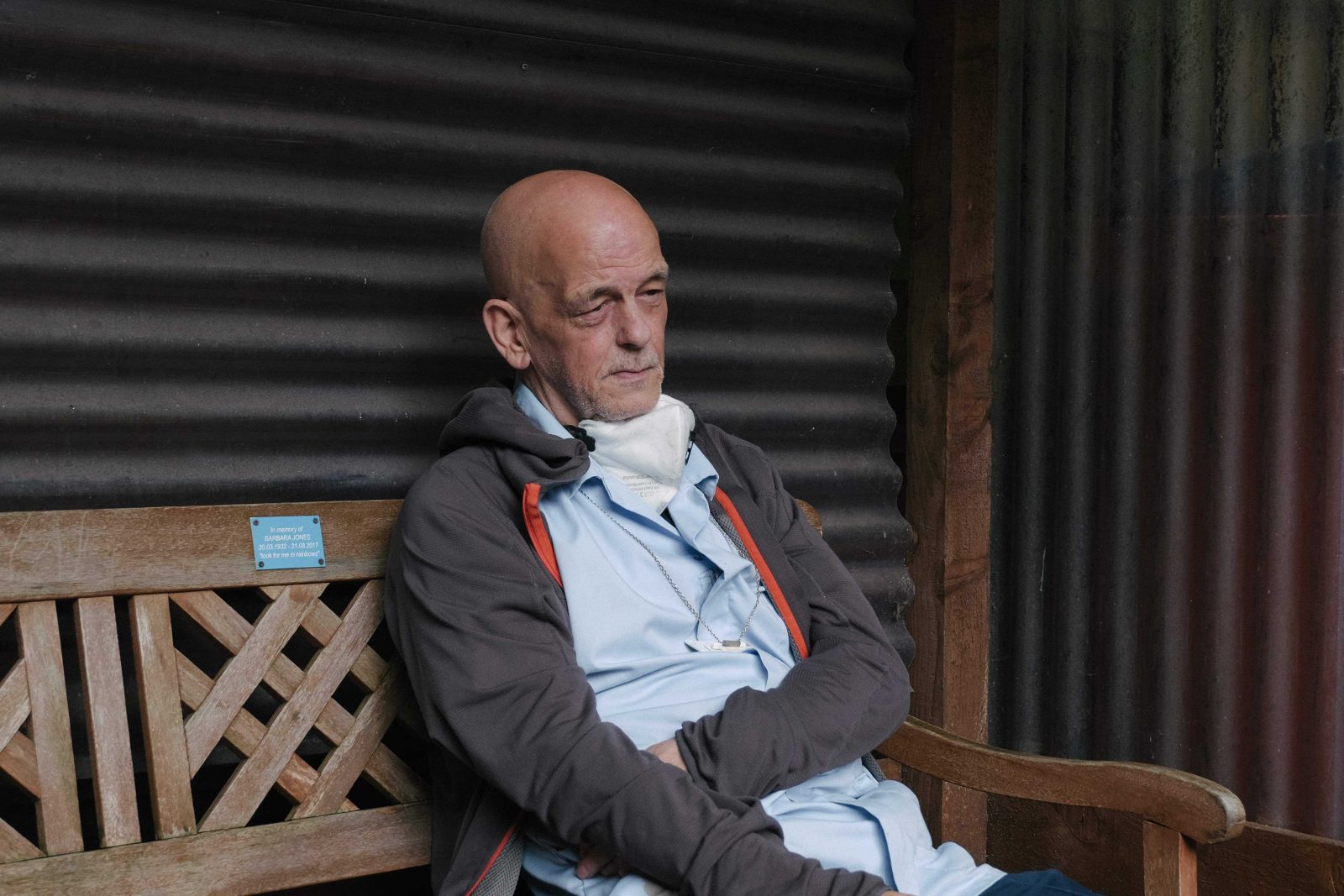 A bald middle aged man is sat on a wooden bench with a face mask pulled down so that it is sitting around his neck. He wears a light blue shirt and a grey hoodie, he looks tired, thoughtful and maybe even slightly angry.