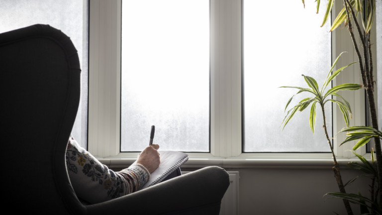 A elderly woman sits in an arm chair facing a window. She is drawing on a piece of paper.