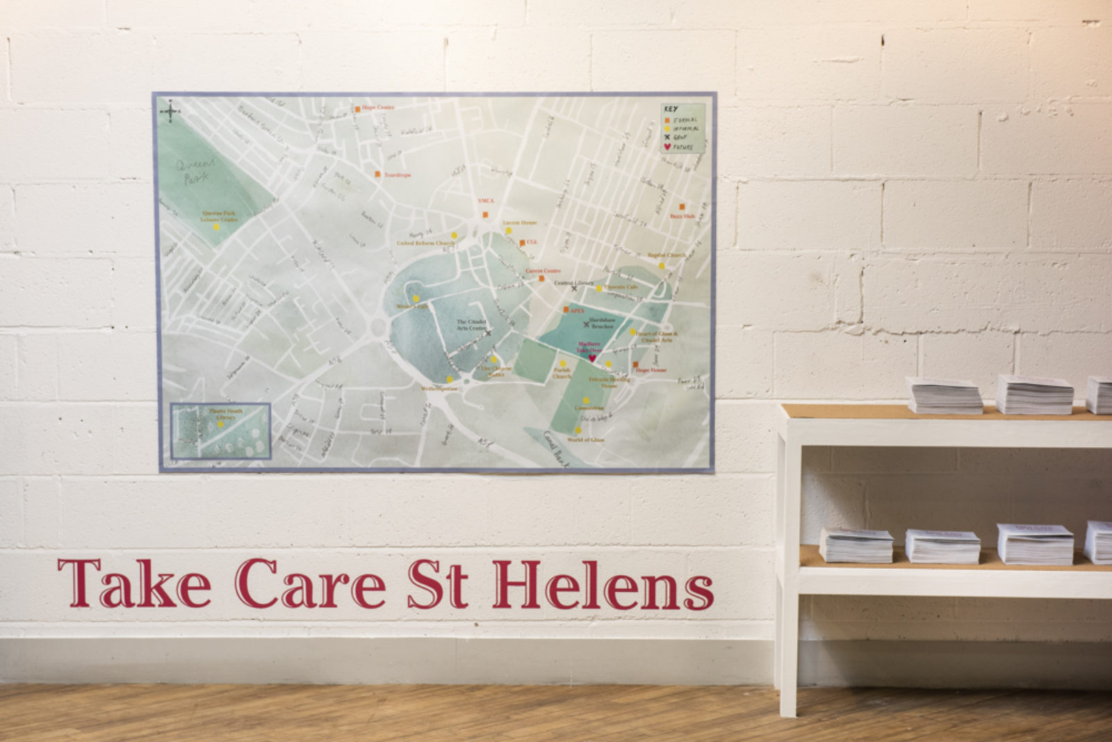 A large map hangs on a wall with a low set of shelves next to it with leaflets on top. Underneath the map the words 'Take Care St Helens' are printed in large red letters.