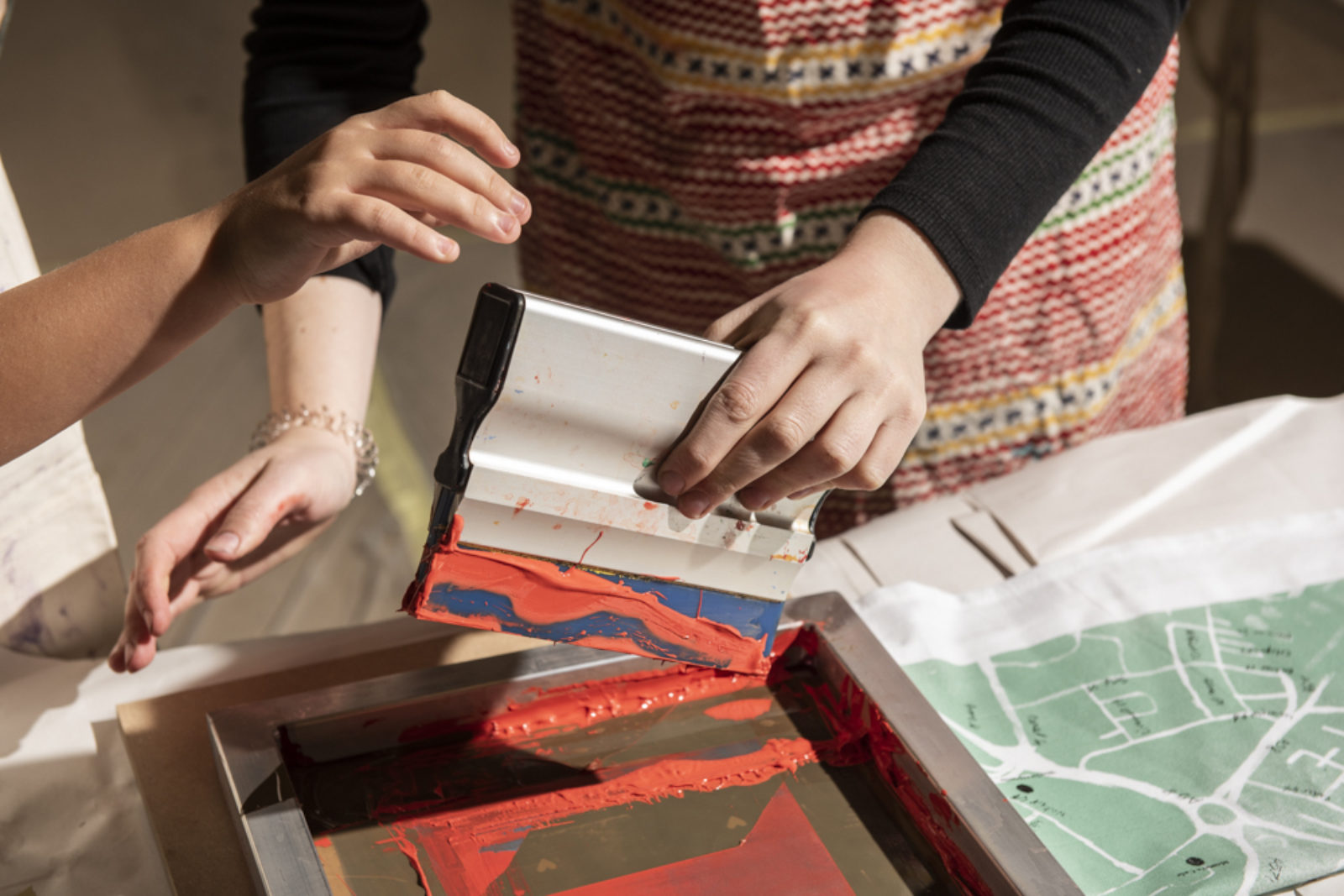 Two pairs of hands are screen printing. Next to the screen a piece of fabric with a map printed on it.