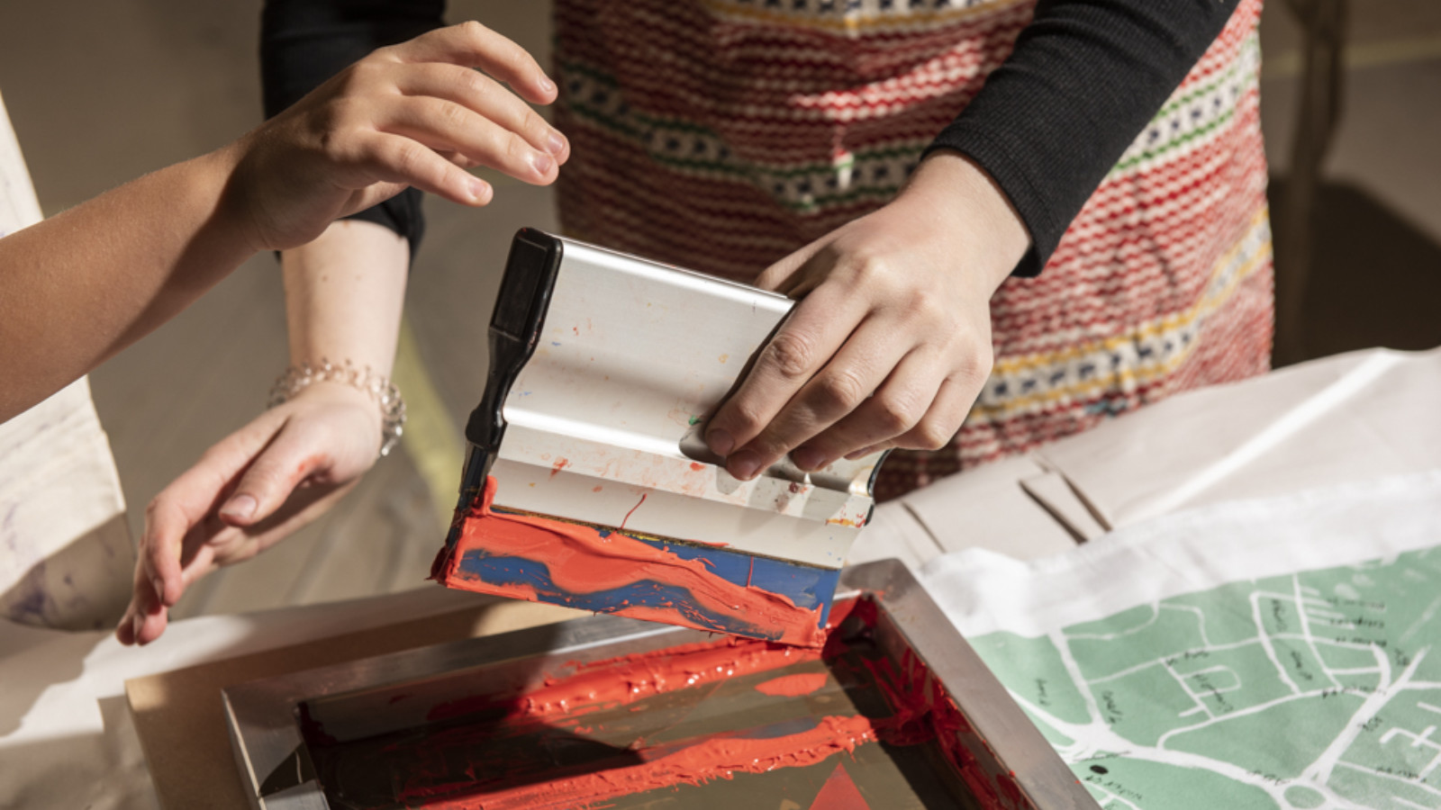 Two pairs of hands are screen printing. Next to the screen a piece of fabric with a map printed on it.