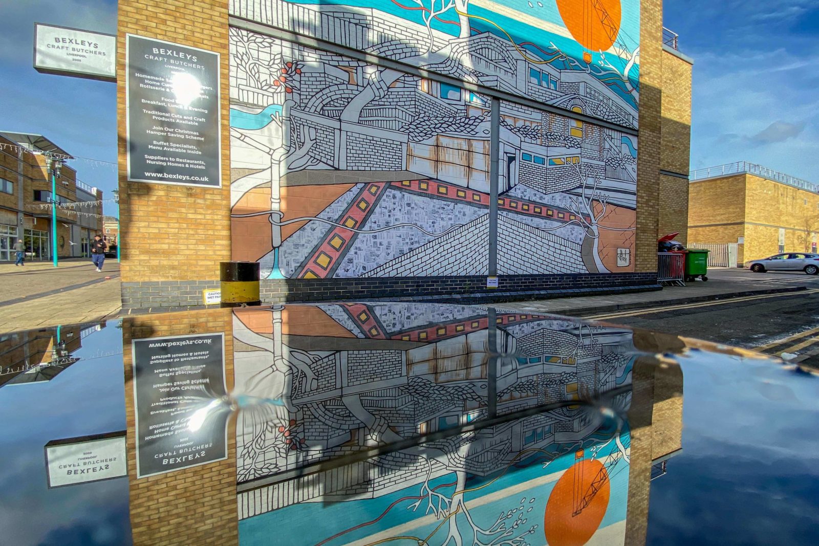 The mural is photographed on a sunny day, the puddle on the ground below it shows its reflection.