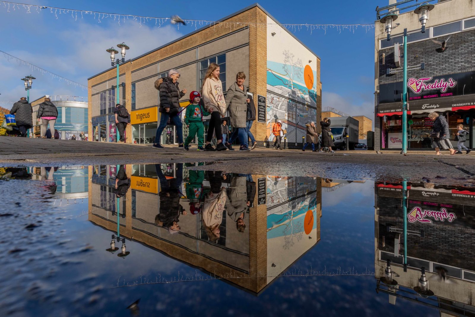 a family walks past the mural on Huyton High Street, their reflections are shown in a puddle below them, and the mural's reflection is behind them.