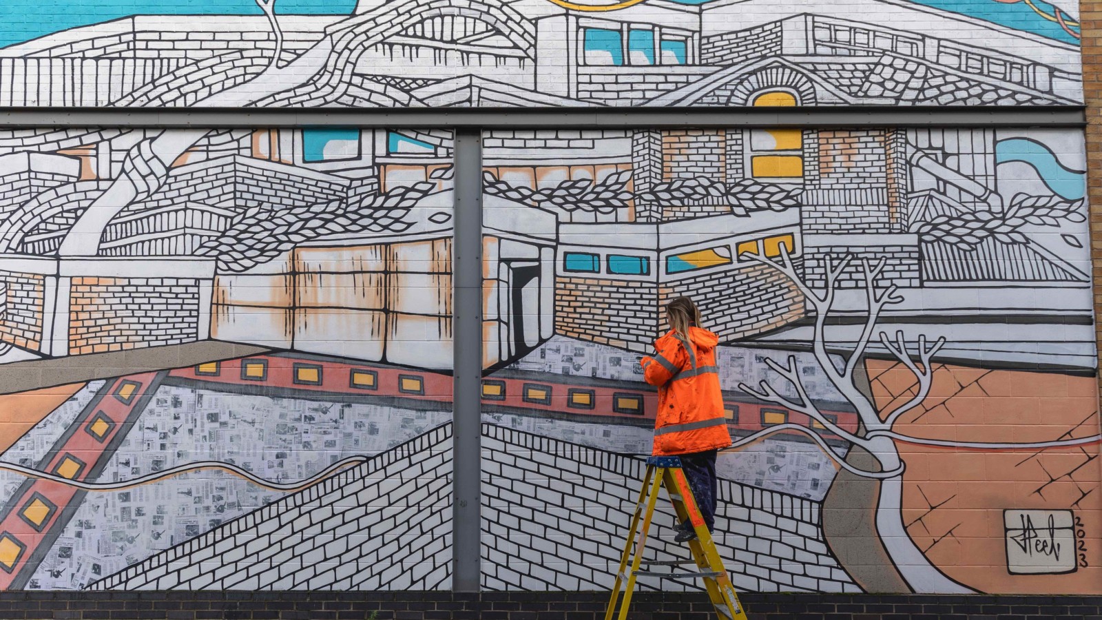Jo Peel is photographed from behind wearing an orange High Vis jacket, stood on a ladder, painting the mural.