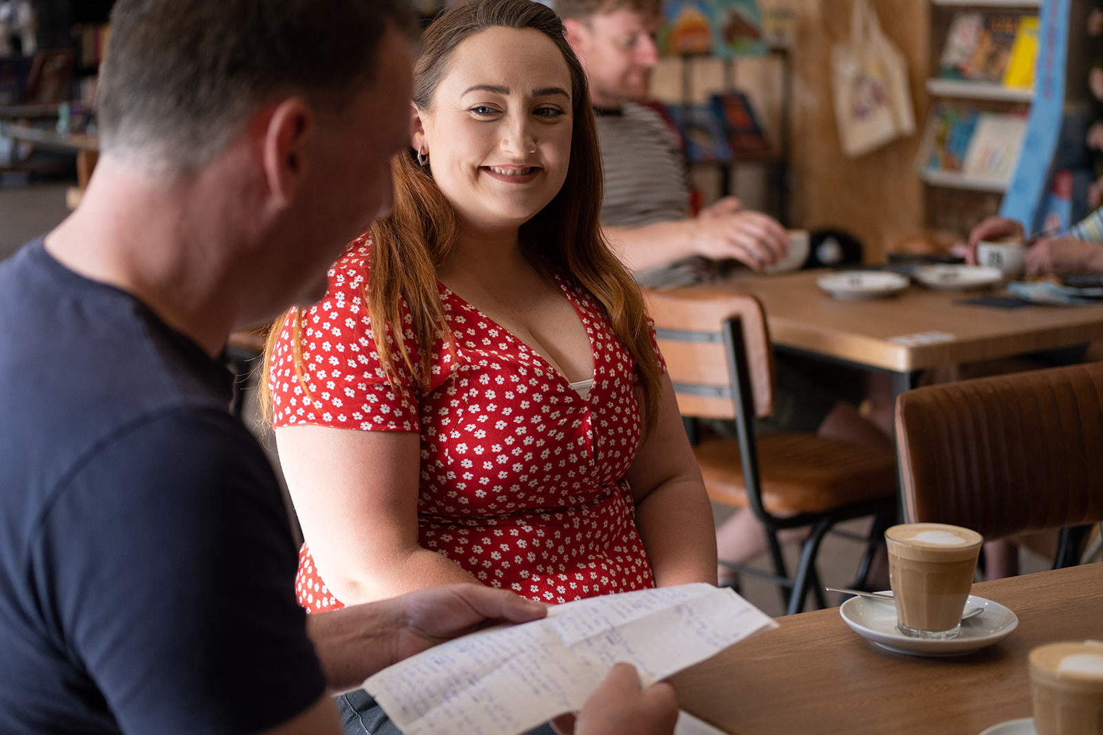 A brunette woman in a red and white polka dot dress smiles at a man next to her on the table in a café as he reads out his answers from a sheet of paper.