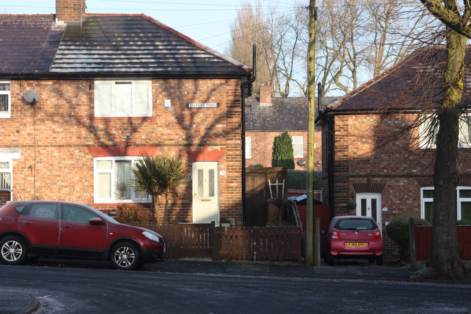 Dickens Road sign is fixed on a semi-detached red brick house with a red car parked in front of it. The roof is frosty and the sky is blue.
