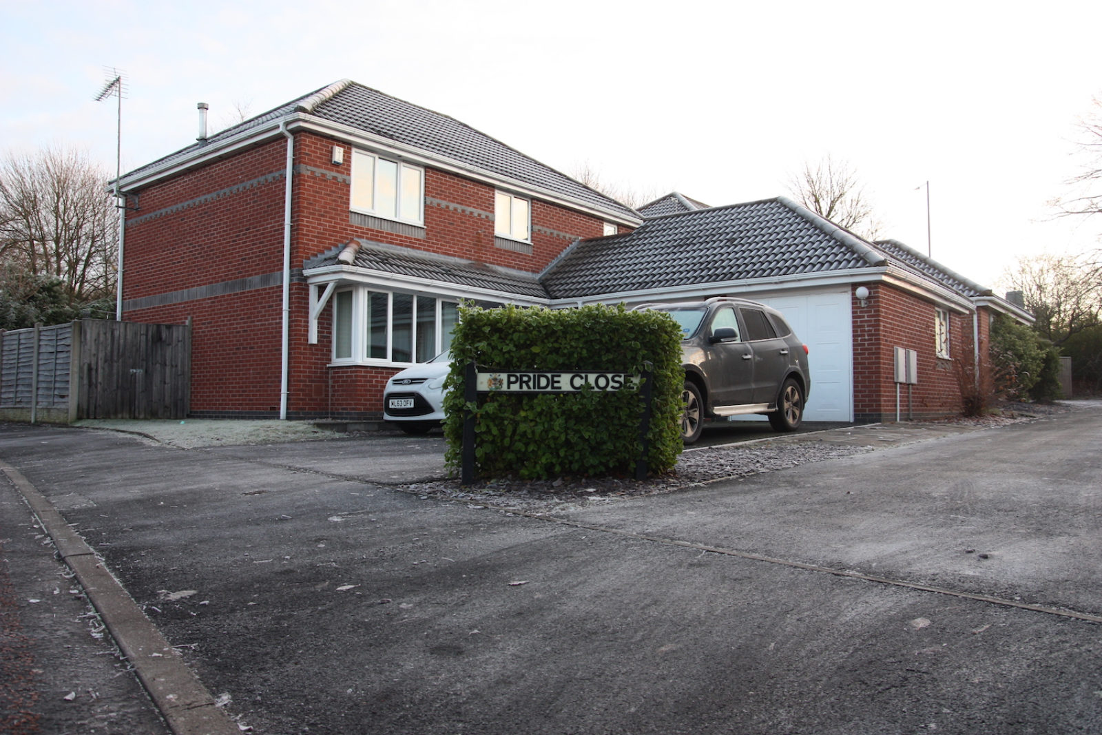 Pride Close sign is inside a hedge on the corner of a driveway of a red brick house with a garage and 2 cars.