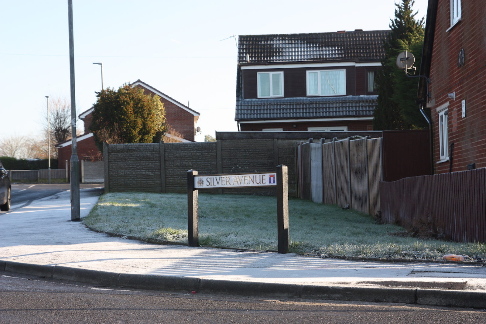 Silver avenue sign is on a street corner with a small patch of grass, fences and red brick houses behind it. The pavement, road and grass are all covered with a tick layer of frost.