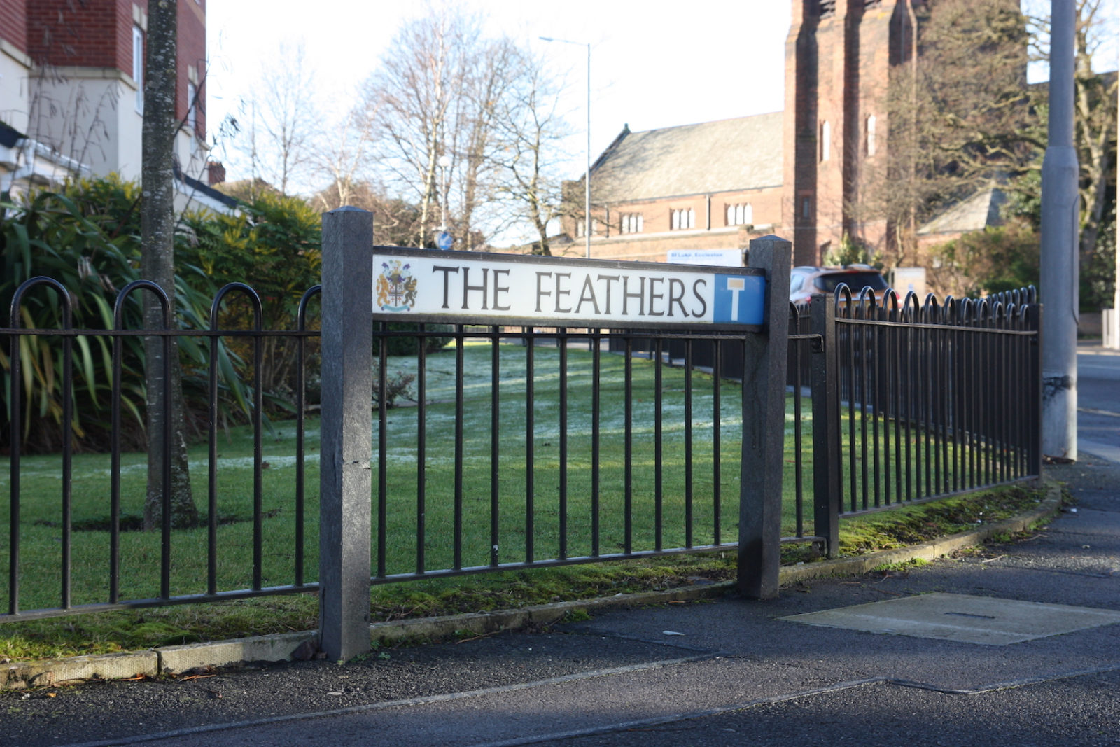 Sign for The Feathers is in front of a short black railing with townhouses, foliage and a church in the background