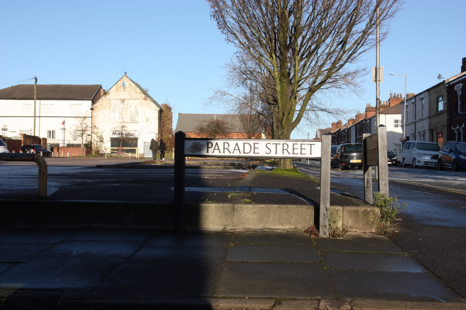 Parade Street Sign stands in front of an empty car park, there is an abandoned looking building in the background.