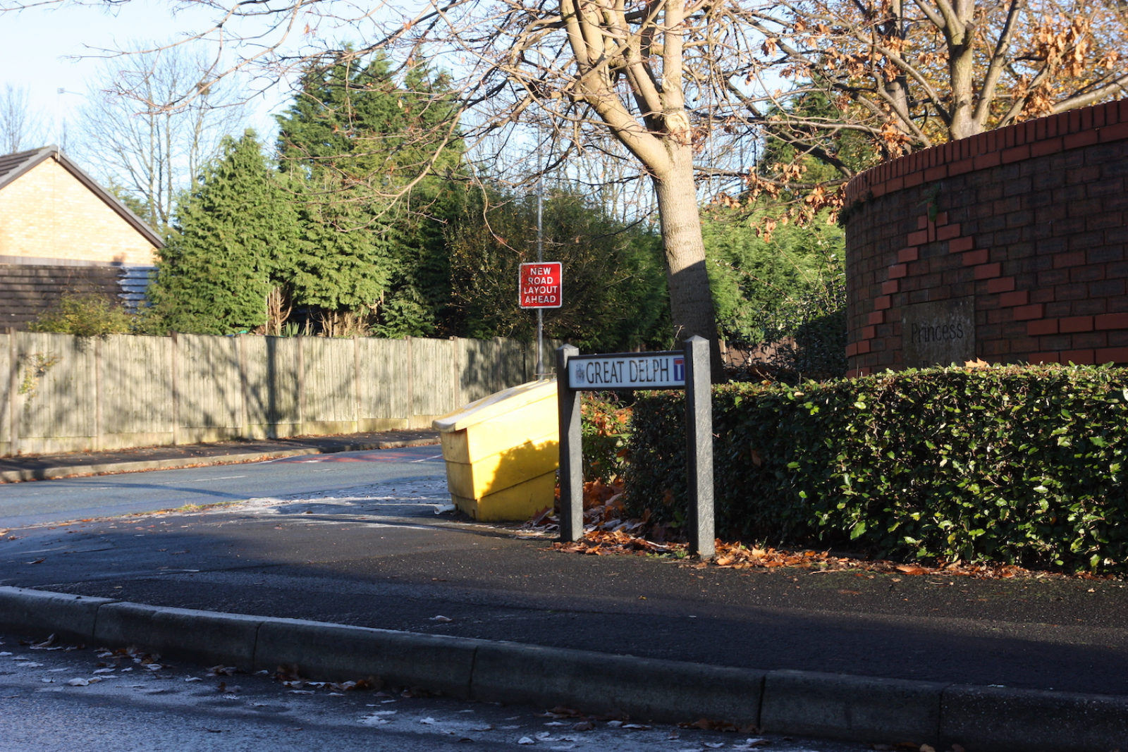 Street sign for 'Great Delph', is on a street corner in front of a short hedge.