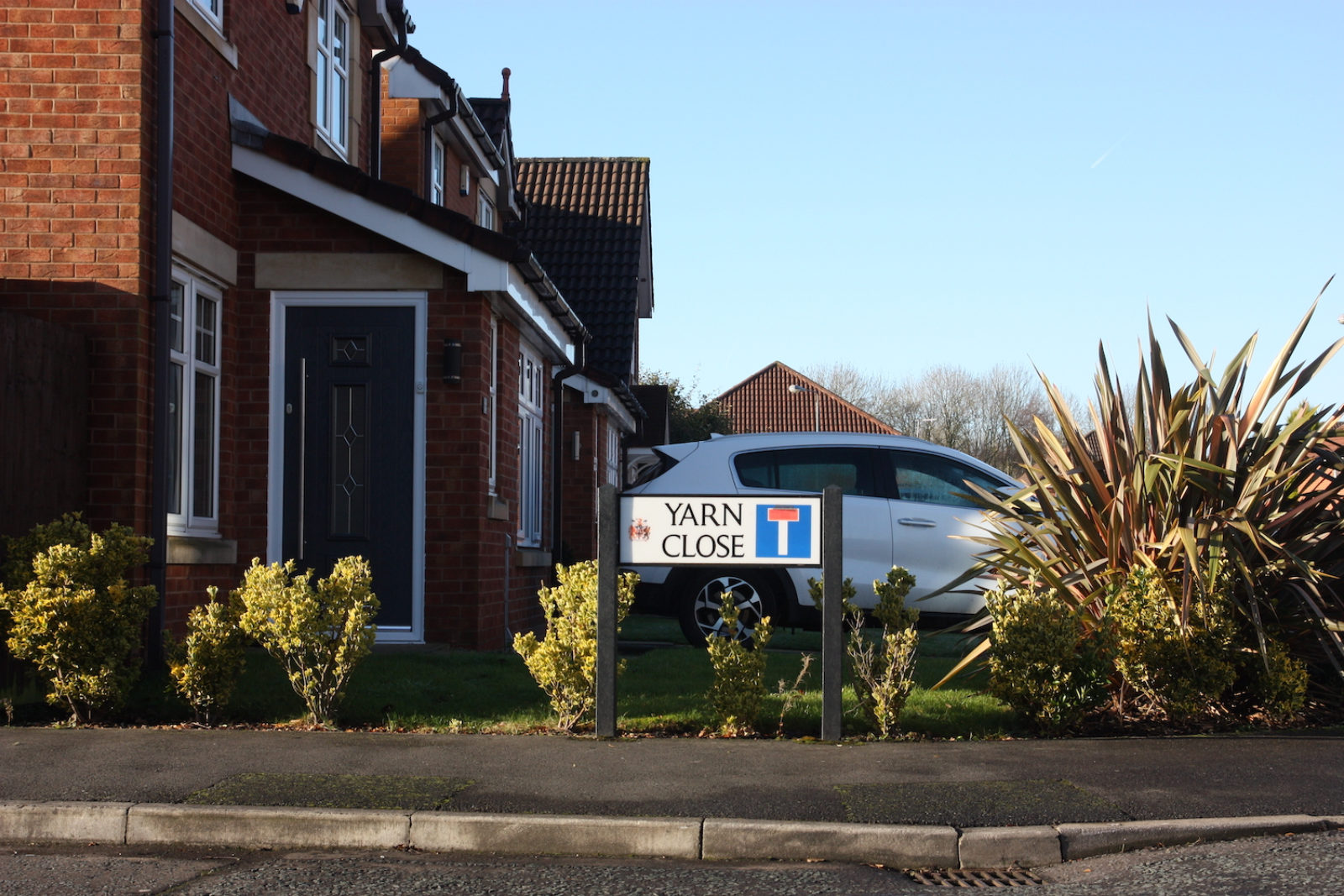 Yarn Close Street Sign is in front of a shite car, bushes and a red brick house with a porch.