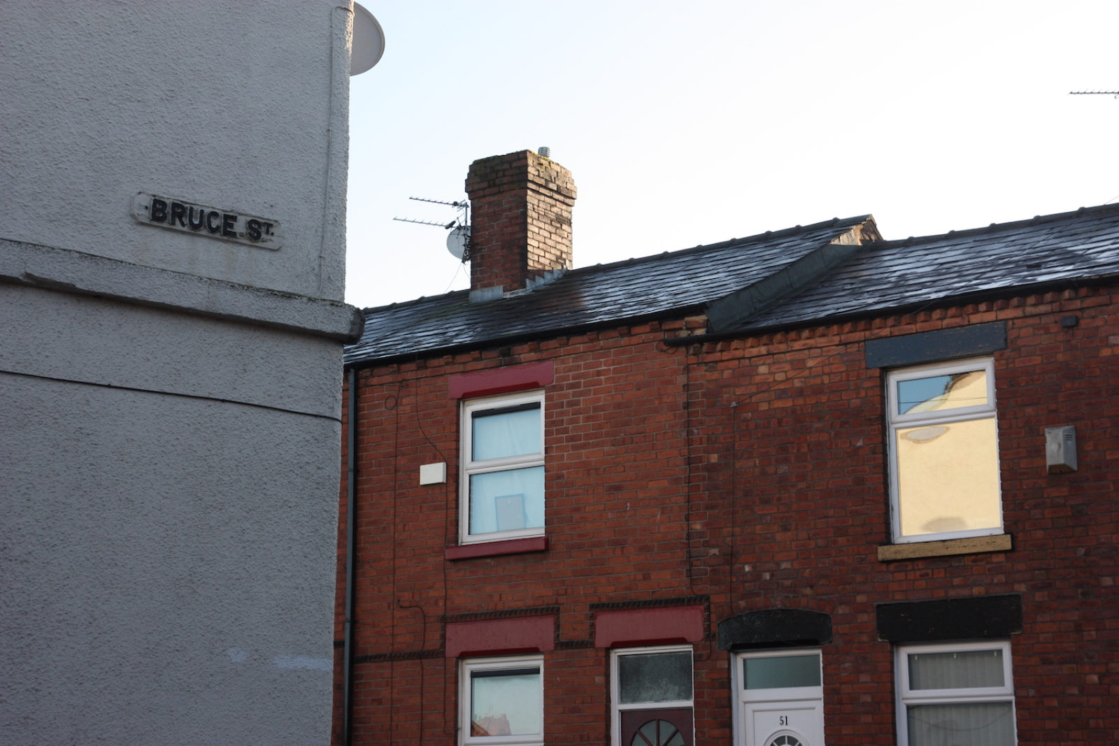 Bruce St sign is on a white wall on the corner of a house, adjacent to red terrace houses.