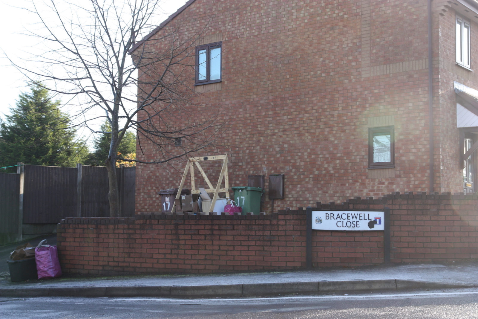 Bracewell Close sign stands in front of a red brick wall, behind the wall is a red brick house with wheels bins in front of it.