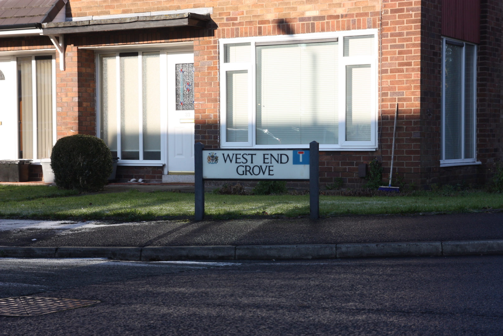 West End Grove sign stands in front of a front garden of a red brick houser with a white front door with a stained glass panel on it, and large white framed windows.