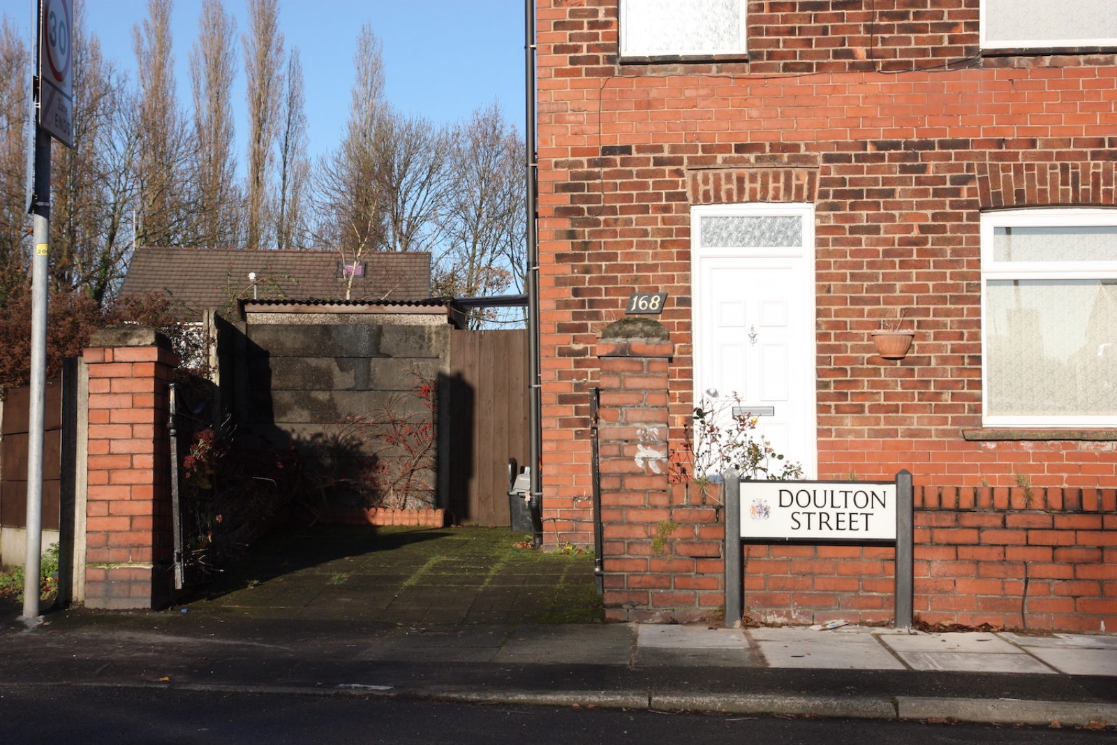 Doulton Street sign is in front of a red brick house, there are bare trees and blue sky in the background.