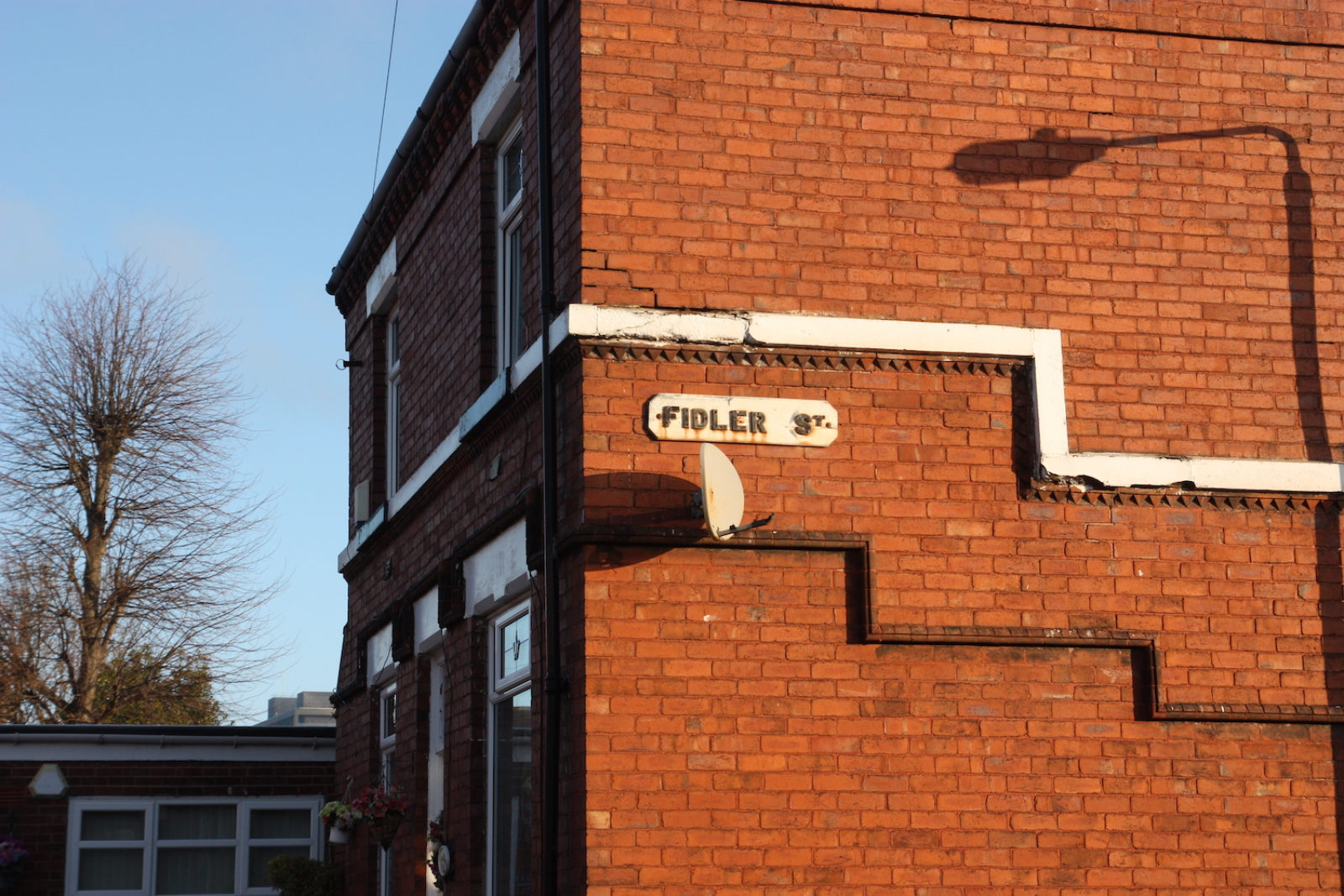 Fidler St. sign is old fashioned and attached to the side of a red brick house with the shadow of a lamppost cast onto it.