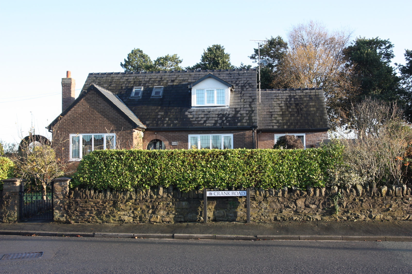 Crank Road sign is in front of a stone wall with a hedge and in front of a red brick house.