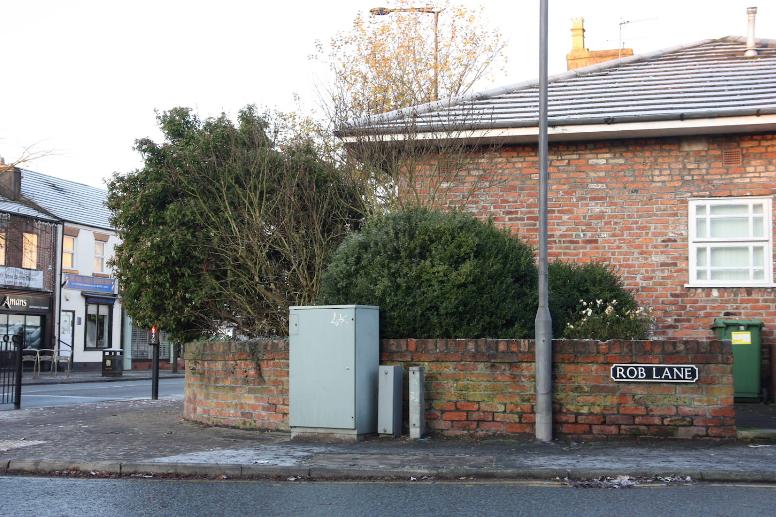 Rob Lane sign is black with white letters and is fixed to a short mossy red brick wall in front of a bungalow.