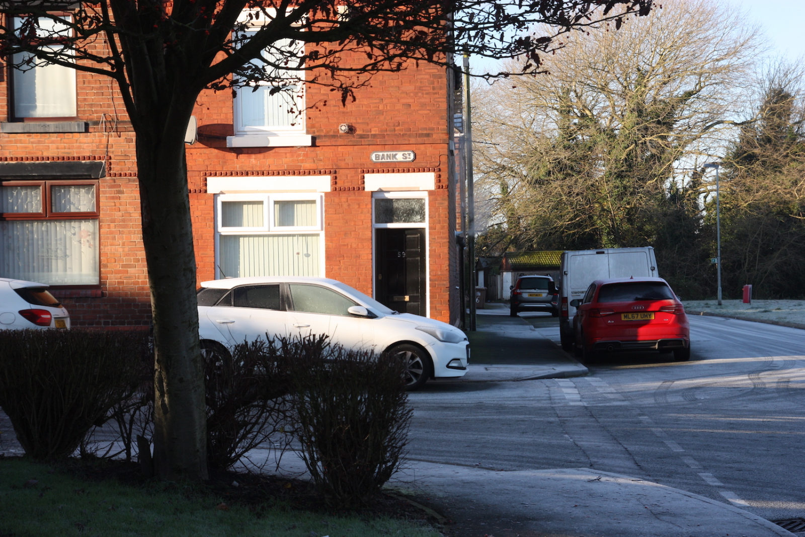 Bank St sign is on the corner of a red brick house above the front door, there are cars parked on the street that are covered in frost.