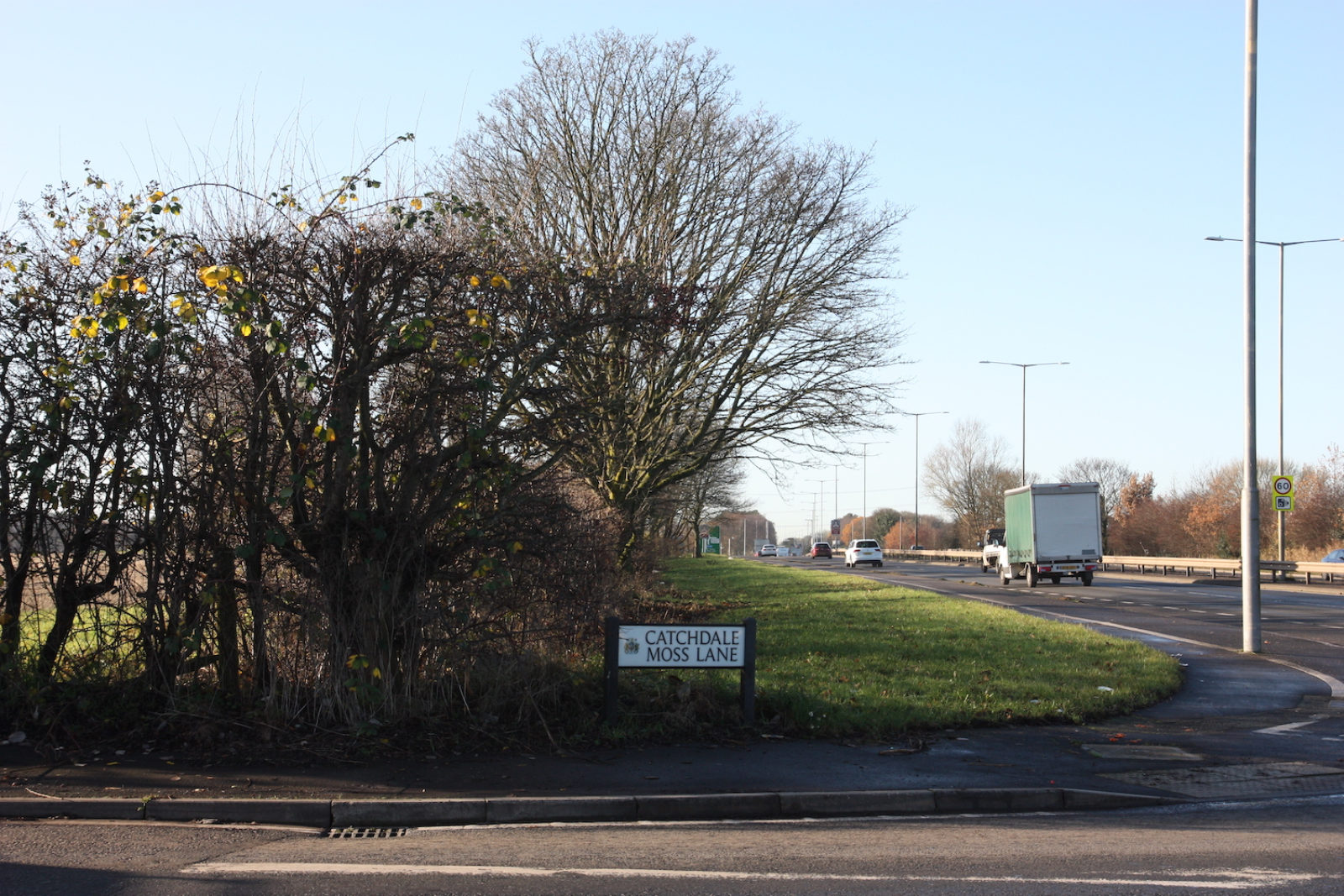 Catchdale Moss Lane sign sits on the corner of a road that leads on to a dual carriageway where there is a green lorry and other cars.