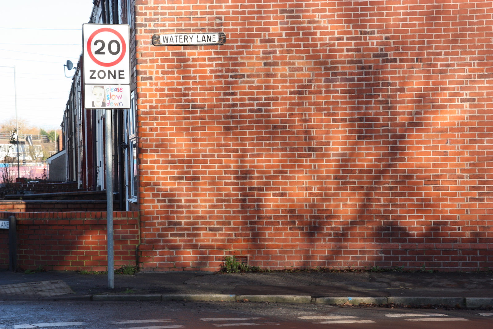 Watery Lane street sign is fitted to the side of a red brick end-terrace house. In front of it is a 20mph zone sign.
