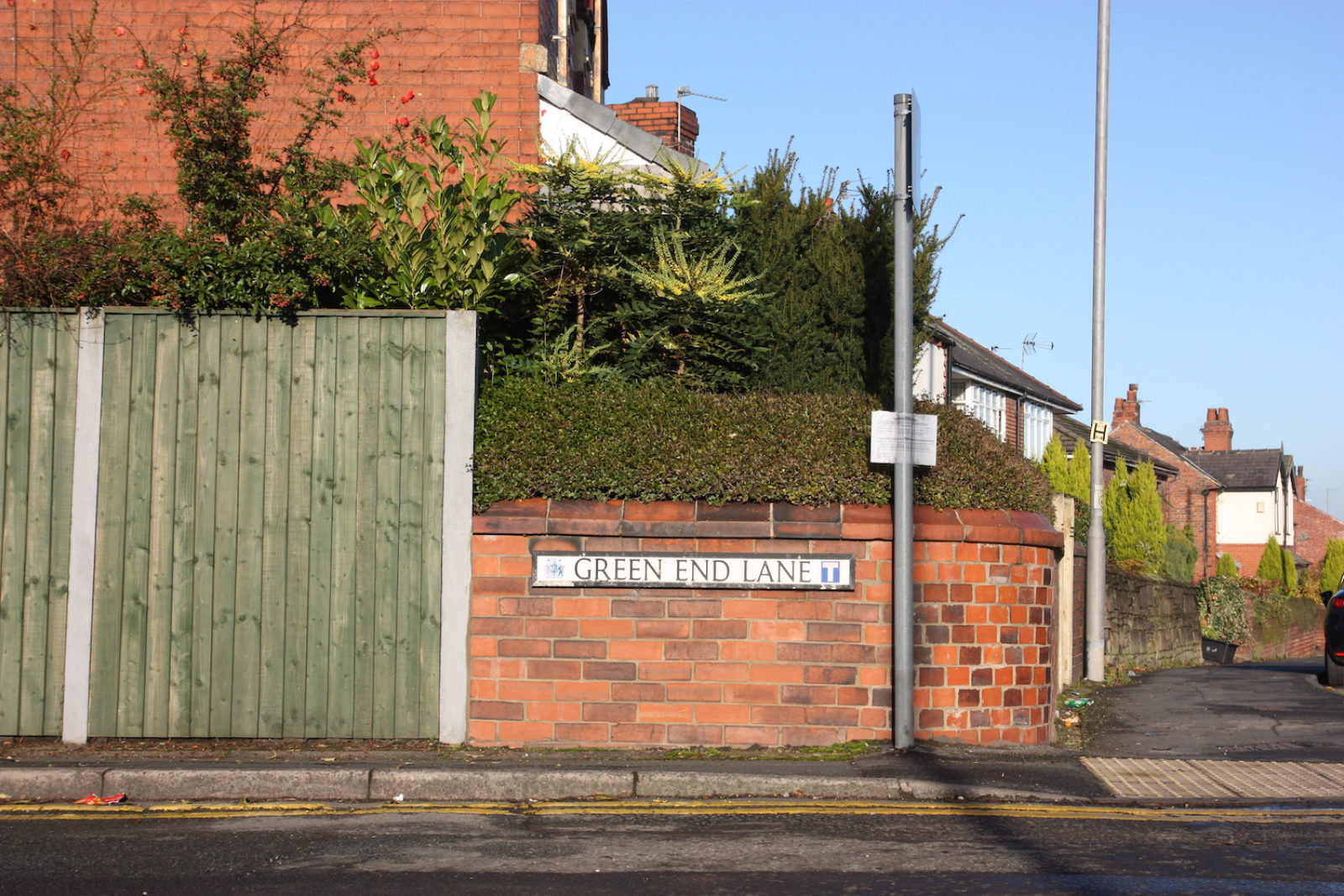 Green end lane sign is on a rounded short brick wall with hedges and foliage behind it. In the background are suburban houses and the sky is blue.