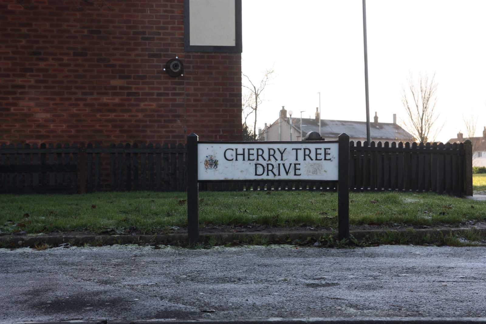 Cherry tree drive is in front of a patch of grass and a red brick wall and the road in front of it is covered in frost.