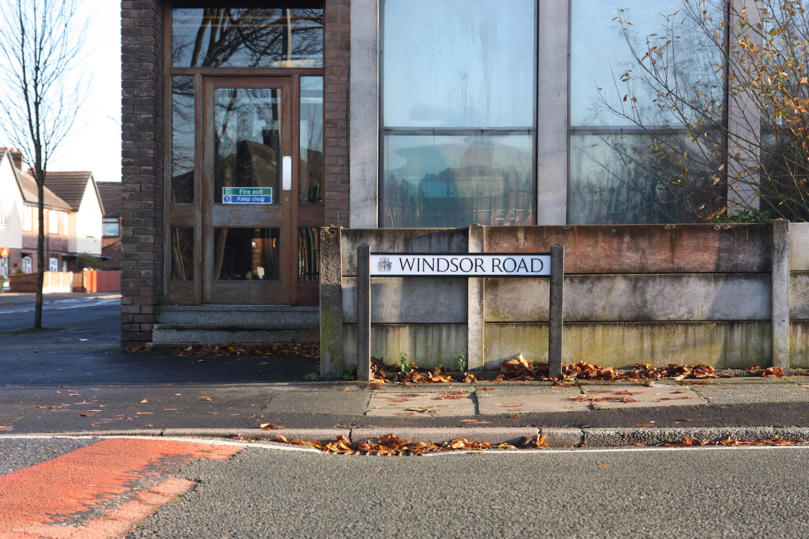 Windsor Road sign iz in front of a mossy concrete wall and a building with large windows and a fire exit. There are autumn leaves on the ground and a speed bump on the road in front.