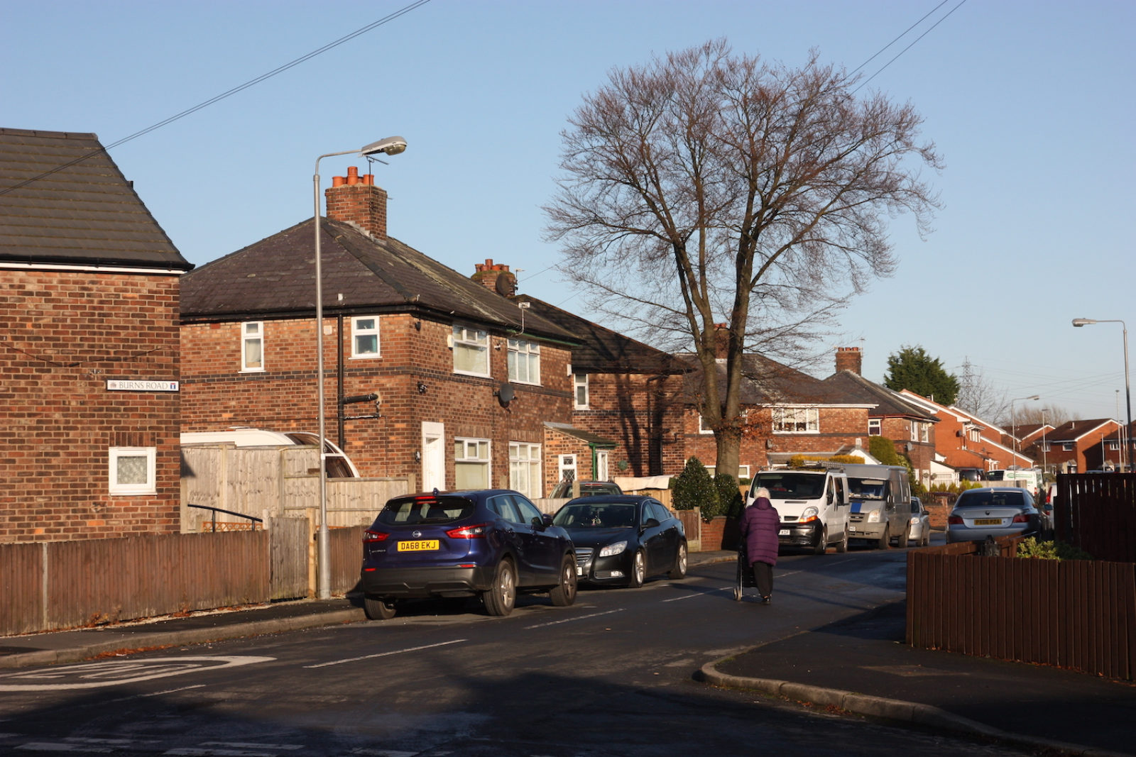 Burns Road sign is on the side of a red brick house, in the background are lots more red brick semi detached houses and an elderly lady walks down the street in a purple coat.