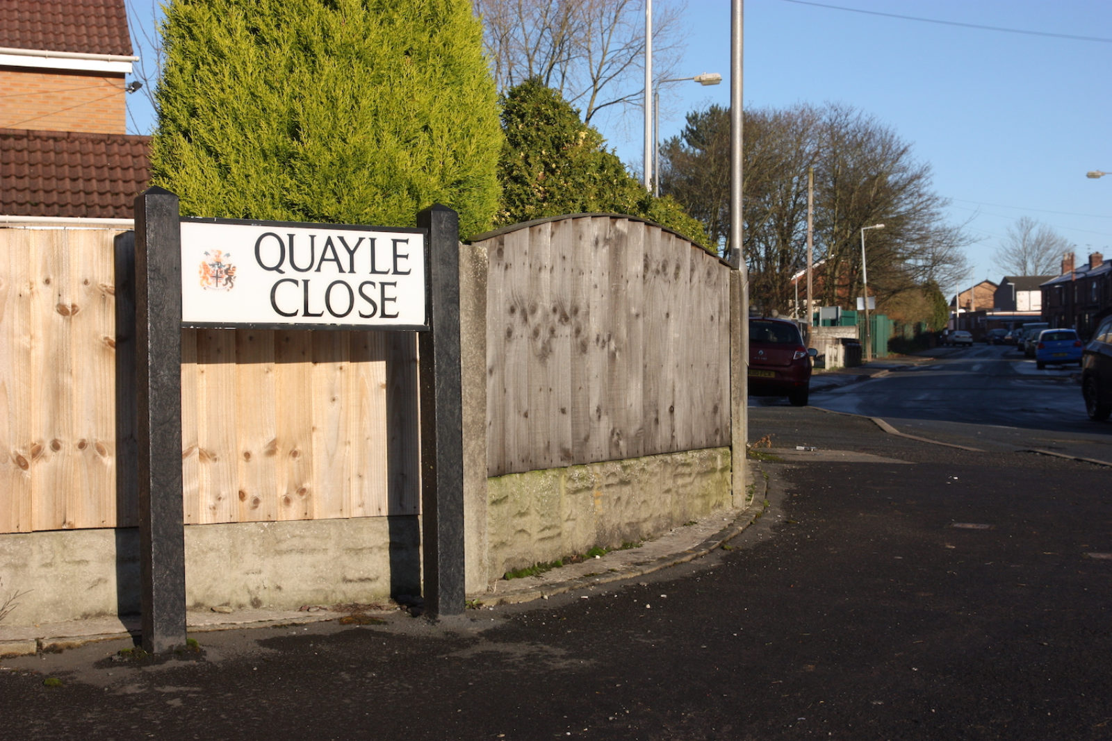 Quayle Close sign stands in front of a wooden fence and hedges.