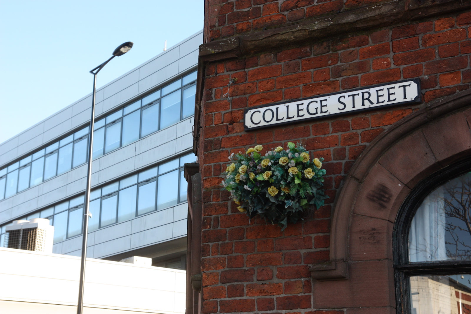 College Street sign is on a red brick wall above an arched window and some hanging flowers.