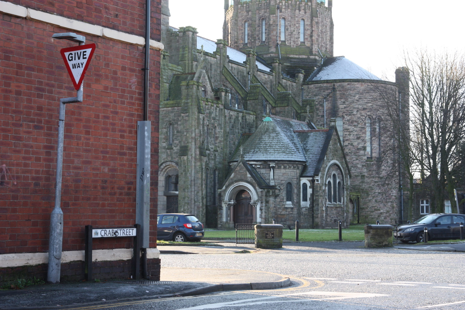 Crab Street Sign is on the corner of a street in front of a red brick wall next to a give way sign, there is a large church in the background on the street adjacent.