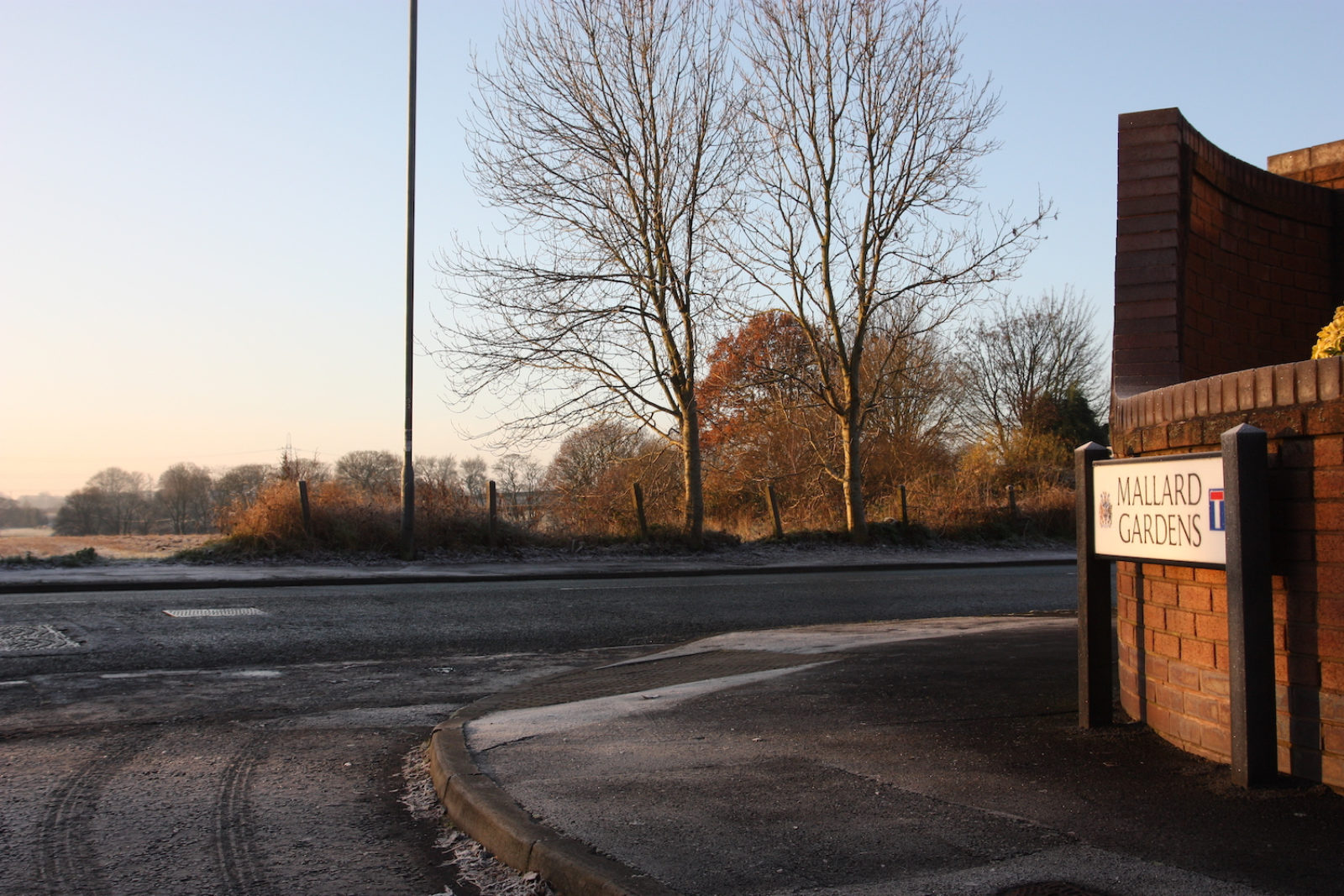 Mallard Gardens sign is on the corner of a street, in the background on the street adjacent are lots of bare trees in front of a frosty field and blue sky.