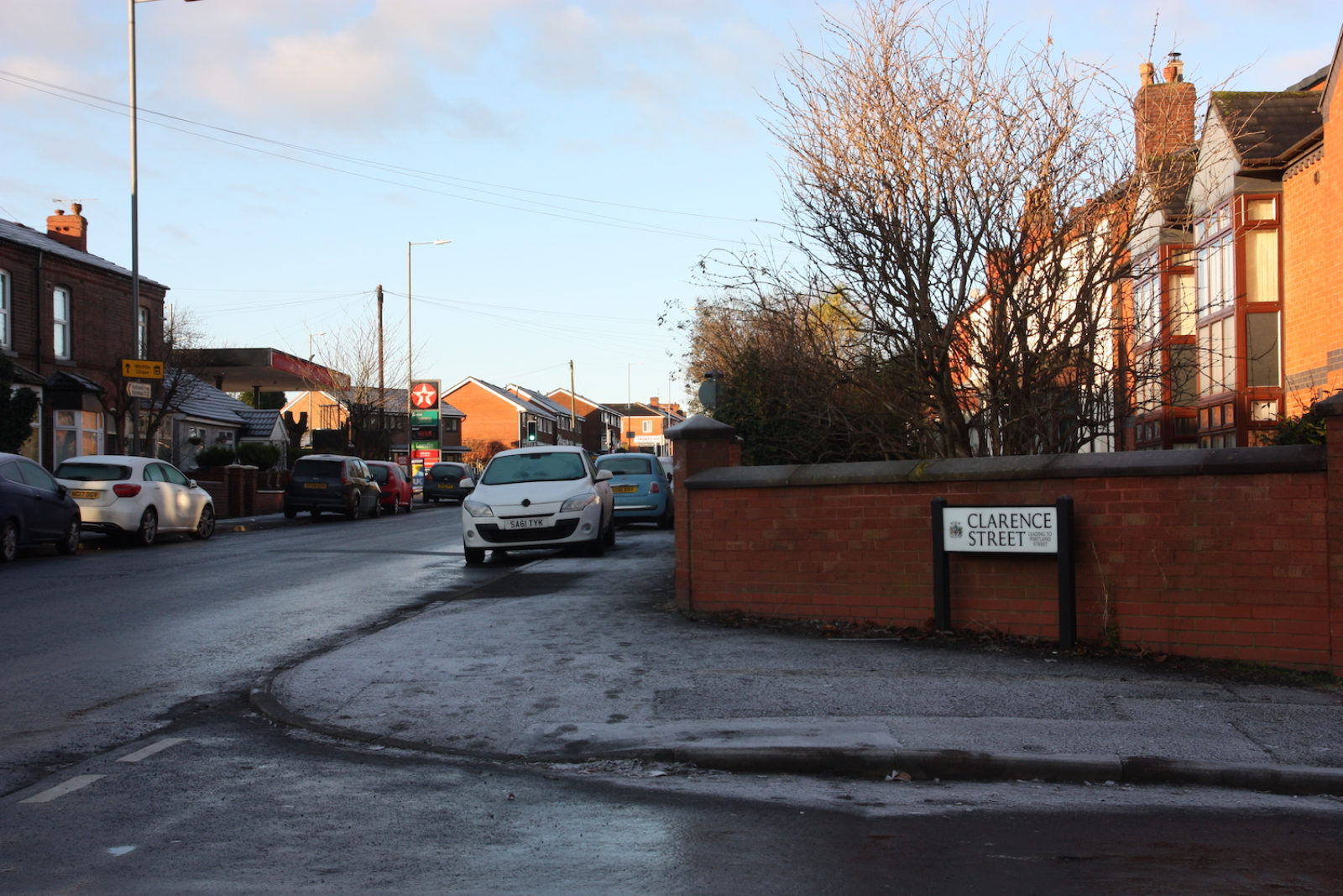 Clerance Street sign is in front of a red brick wall on a street corner, there are houses, trees, parked cars and a petrol station in the background. The sky is blue and the ground is wet.