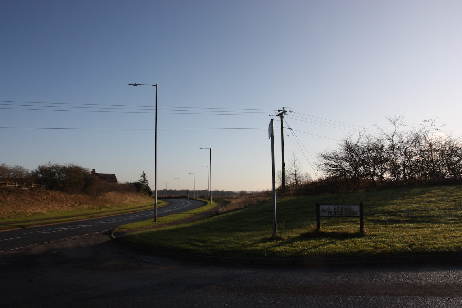 Finger House Lane is on grass on the right of a long winding road with no houses on it.