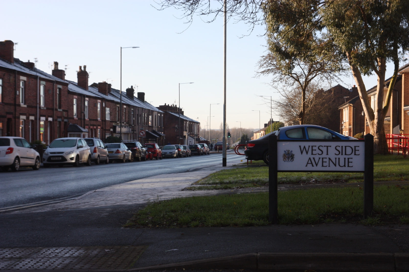 West side avenue sign is on the corner of a road in front of a patch of grass next to as row of terrace houses.