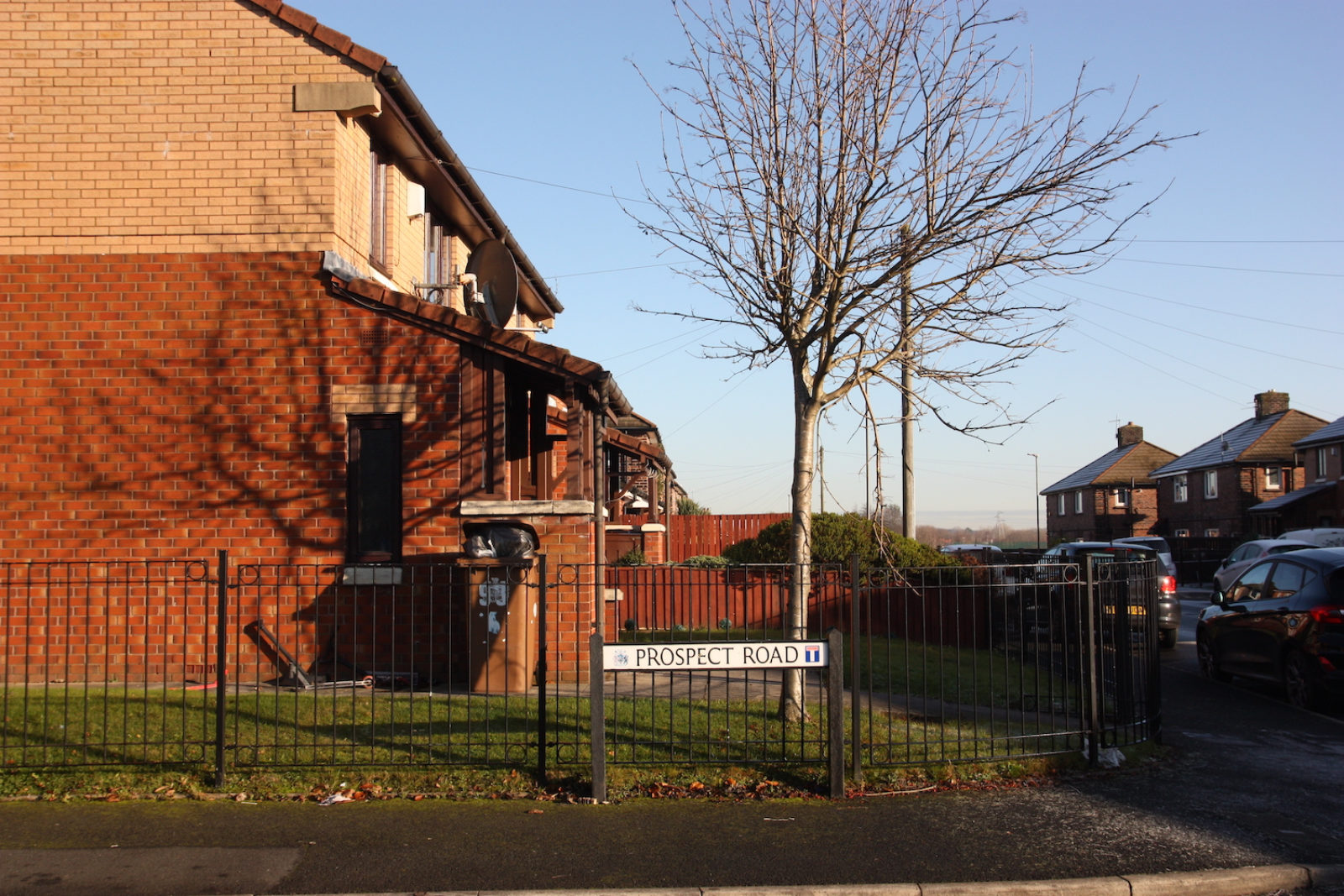 Prospect road sign is in front of small black railings in a suburban area, the sun is low and the sky is blue, there is a bare winter tree behind the street sign.