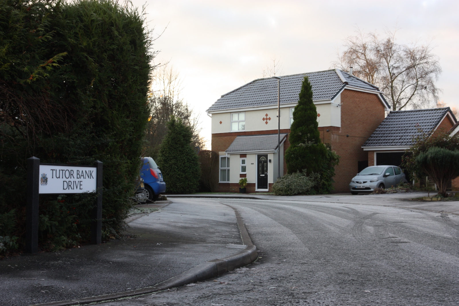 Tutor Bank Drive sign is in front of a hedge on a frosty street with a half white half red brick house in the background.