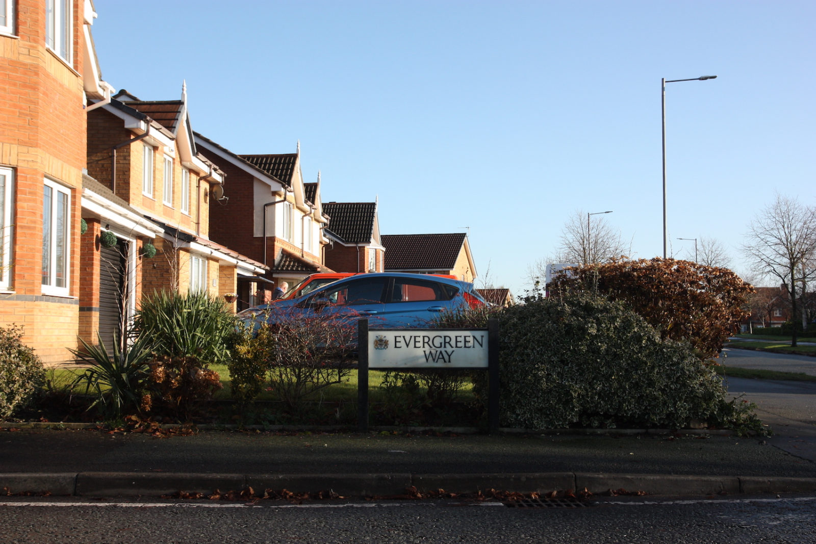 Evergreen Way sign stands in front of a front garden with a blue car parked behind it, there are red brick new build houses in the background and it is sunny.