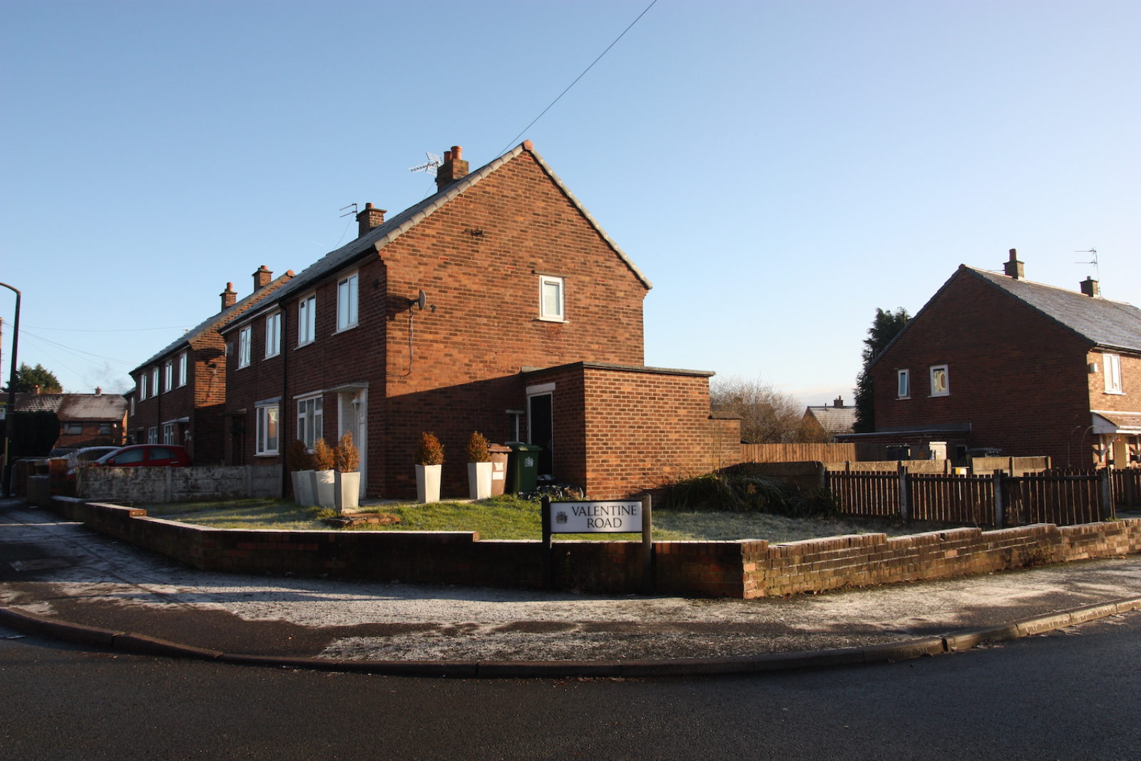 Valentine Road sign is on the corner of a street in front of a front garden with a short wall surrounding it, there are red brick semi-detached houses in the background.