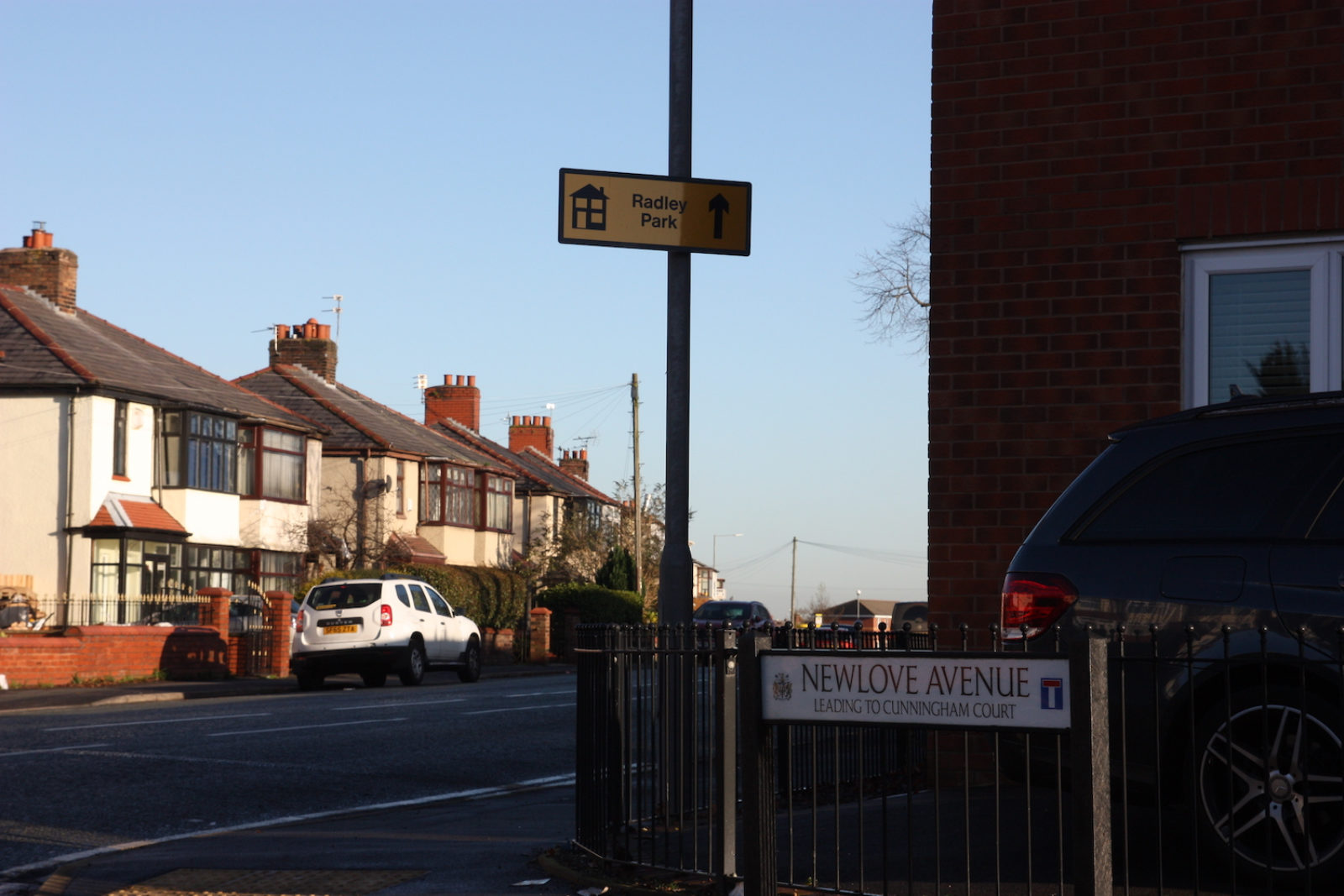 Newlove Avenue street sign is at the end of a road in front of railings, the street adjacent has a sign pointing to Bradley park and white semi detached houses on it.