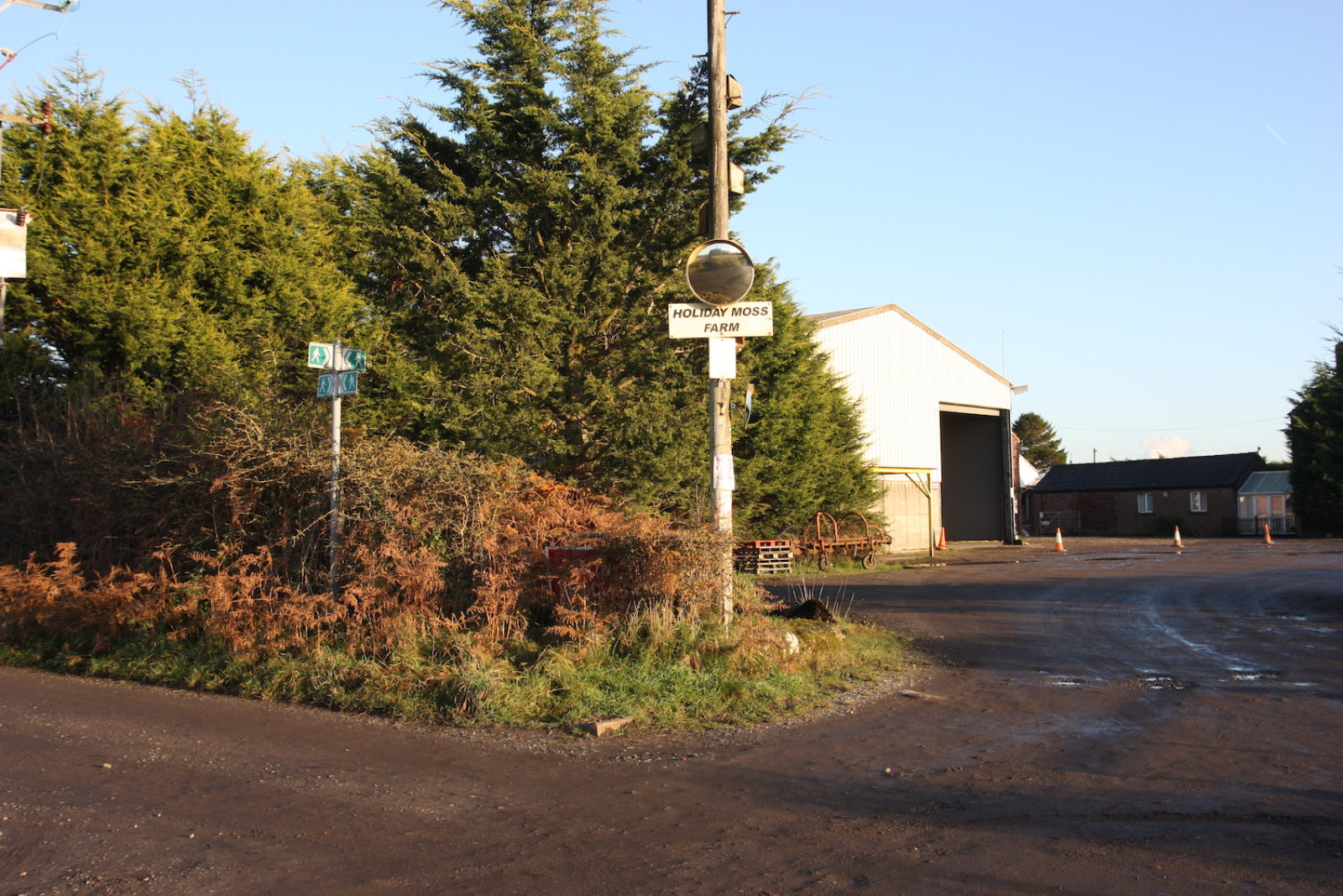Holiday moss farm sign is on a post with a round mirror above it, there are trees and a large industrial building in the background.