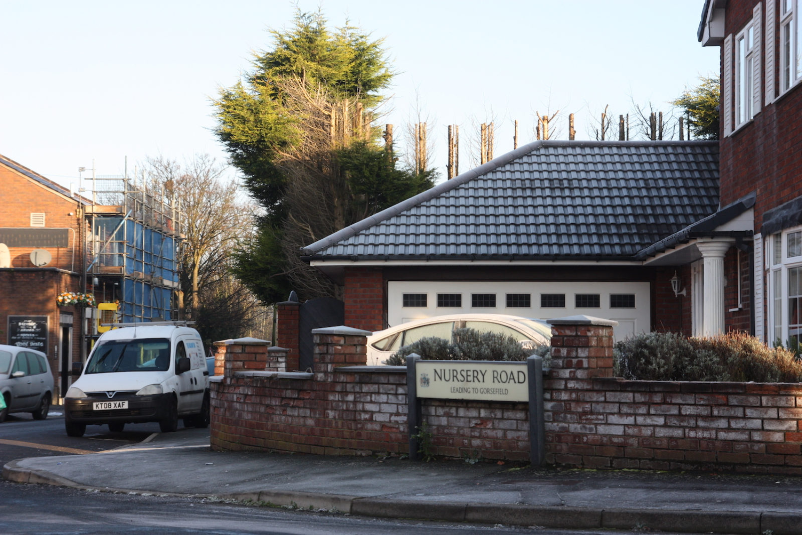 Nursery Road sign stands in front of a short red brick wall, there is a garage with a white door and a frosty roof in the background and parked cars.