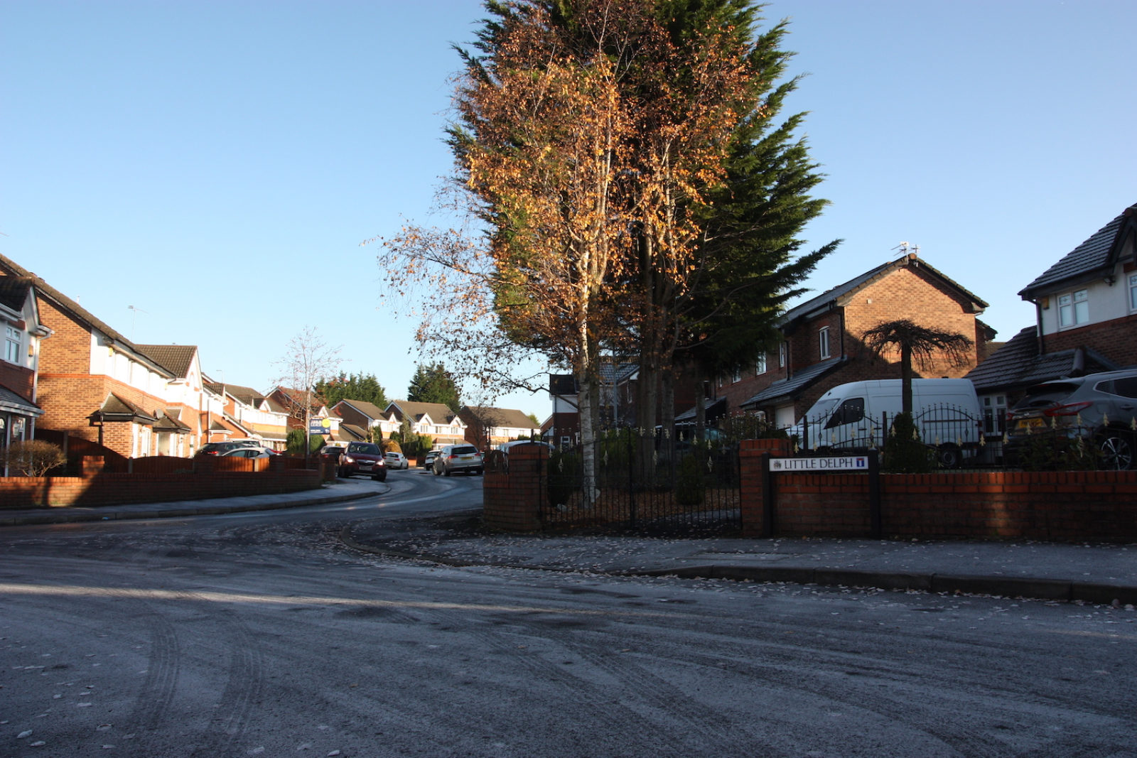Little Delph street sign stands in front of a brick wall and black railings, there is a white van parked in the background and a frosty winding road.