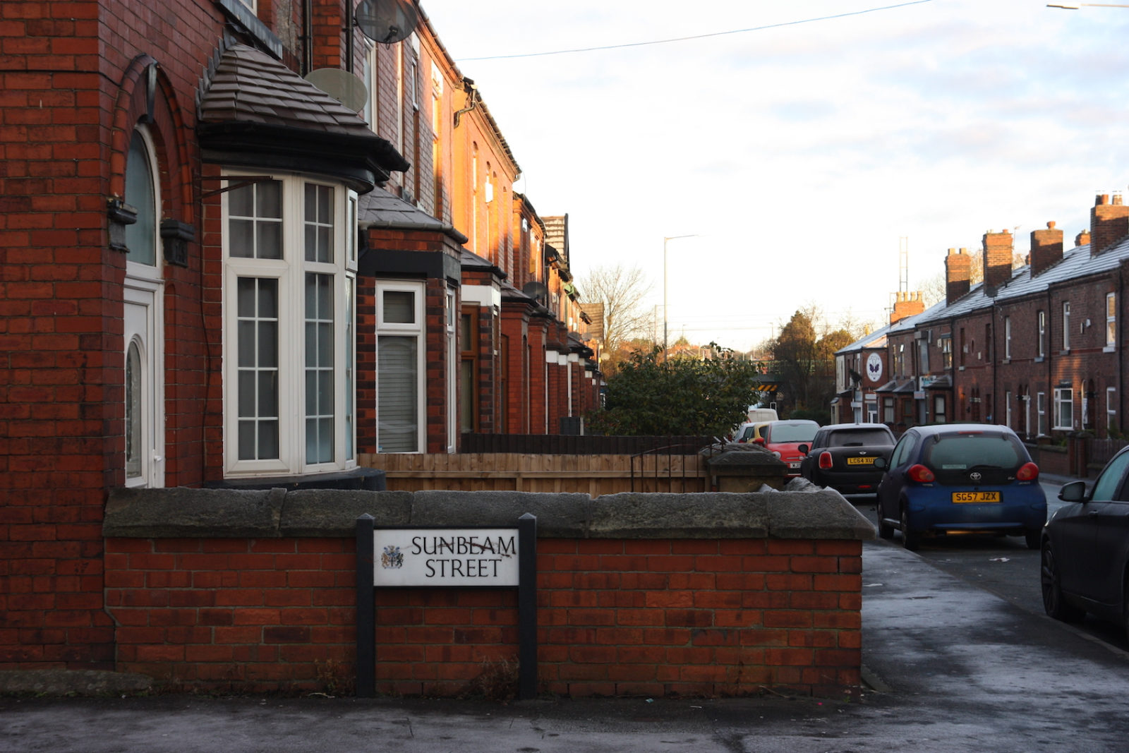 Sunbeam Street sign is in front of a red brick wall of a small front garden at the end of a row of terrace houses.