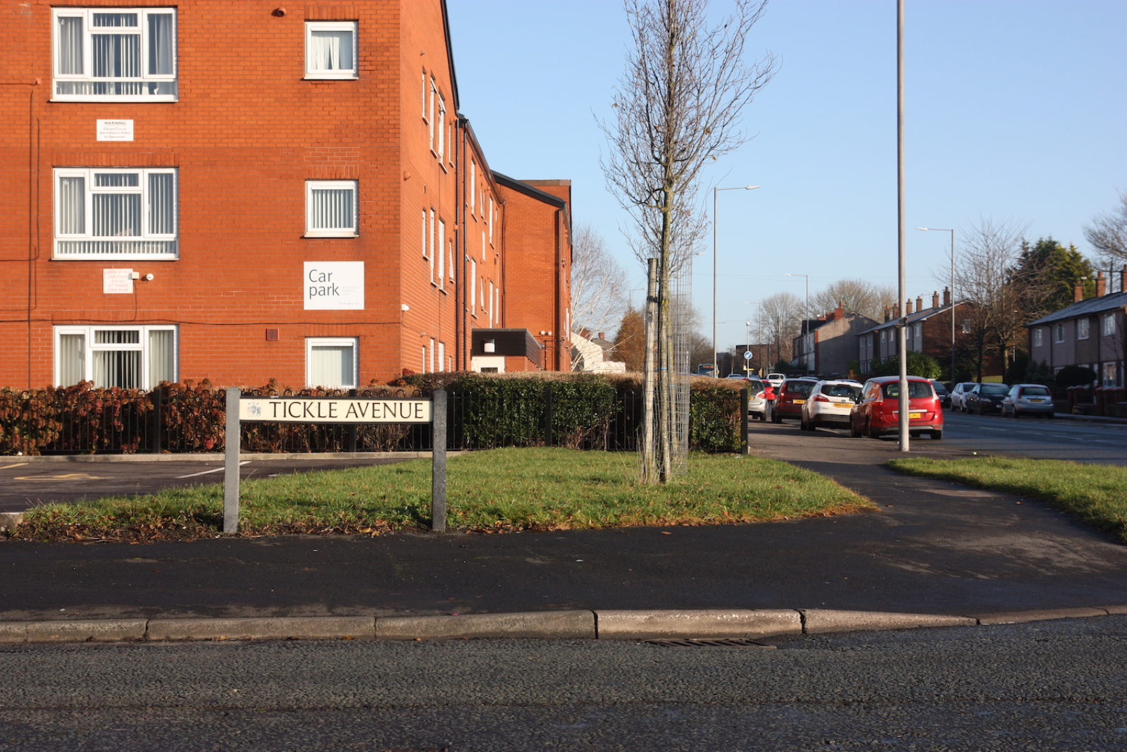 Tickle Avenue sign is on a street corner in front of a patch of grass and a red brick block of flats with a 'Car Park' sign fixed onto it.