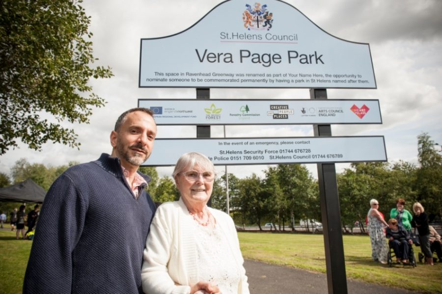 Two people, one older woman and one man, stand with their arms linked in front of a large sign. The sign has a crest at the top and reads 'St Helens Council. Vera Page Park' in large letters.