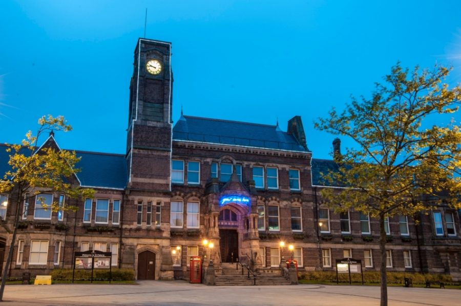 In evening light a large stone building with a clocktower spans the width of the image. The building has a large paved area in front of it with small trees, benches and a phone box. The building has a large wooden door at the top of some stone steps. Above the door is a blue neon sign.